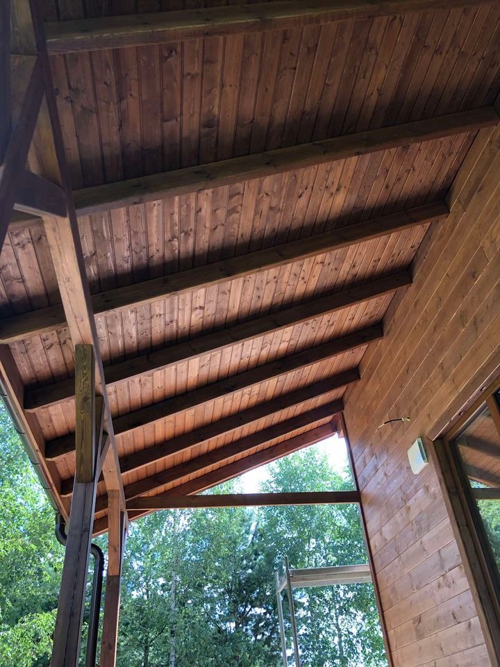 Drewniany sufit z belek i boazerii w altanie z widokiem na zielone drzewa, ciepłe oświetlenie, widoczne łączenia desek.