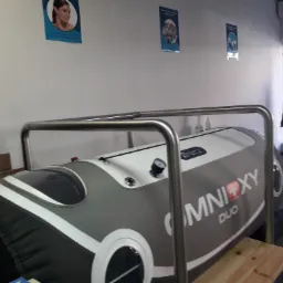 Komora hiperbaryczna Omnioxy Duo w pomieszczeniu z plakatami na ścianie, widoczna platforma wejściowa i krzesło lekarskie w tle.