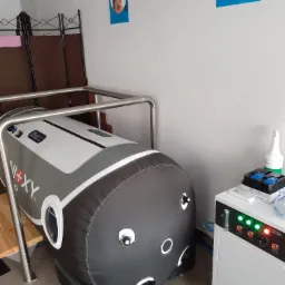 Komora hiperbaryczna w pomieszczeniu zabiegowym, widoczne plakaty informacyjne na ścianie, urządzenie sterujące z panelami kontrolnymi, butelka z płynem dezynfekcyjnym i schodki wejściowe.