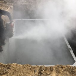 Mat-Instal Mateusz Gładykowski - Cięcie betonu w ziemi, prawdopodobnie otworu, z użyciem piły z chłodzeniem wodnym, generującą parę. Widoczny fragment pracownika i krawędź wykopu.