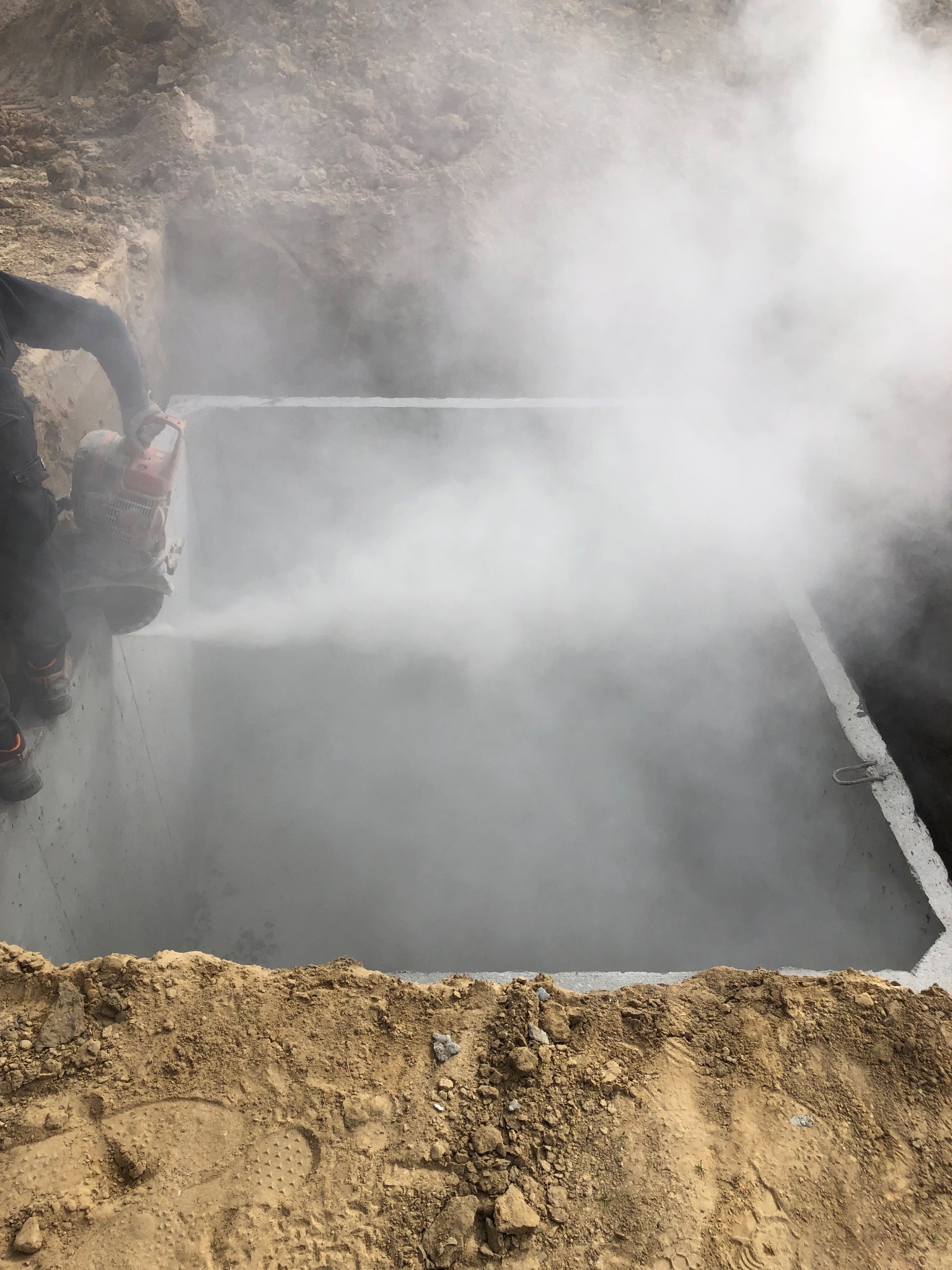 Cięcie betonu w ziemi, prawdopodobnie otworu, z użyciem piły z chłodzeniem wodnym, generującą parę. Widoczny fragment pracownika i krawędź wykopu.