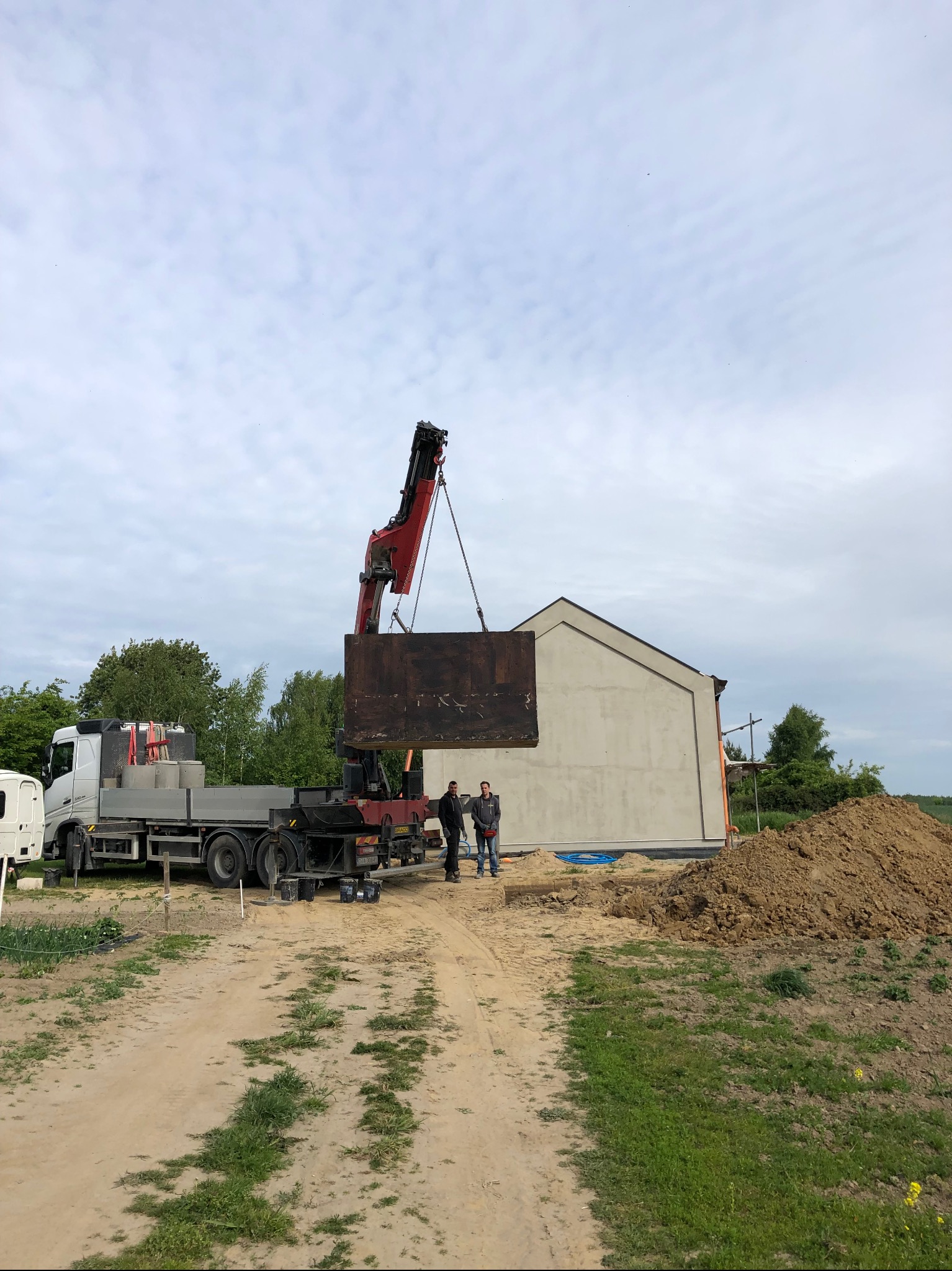 Montaż betonowego szamba przy użyciu dźwigu na placu budowy. Widoczni dwaj pracownicy i ciężarówka z elementami szamba.