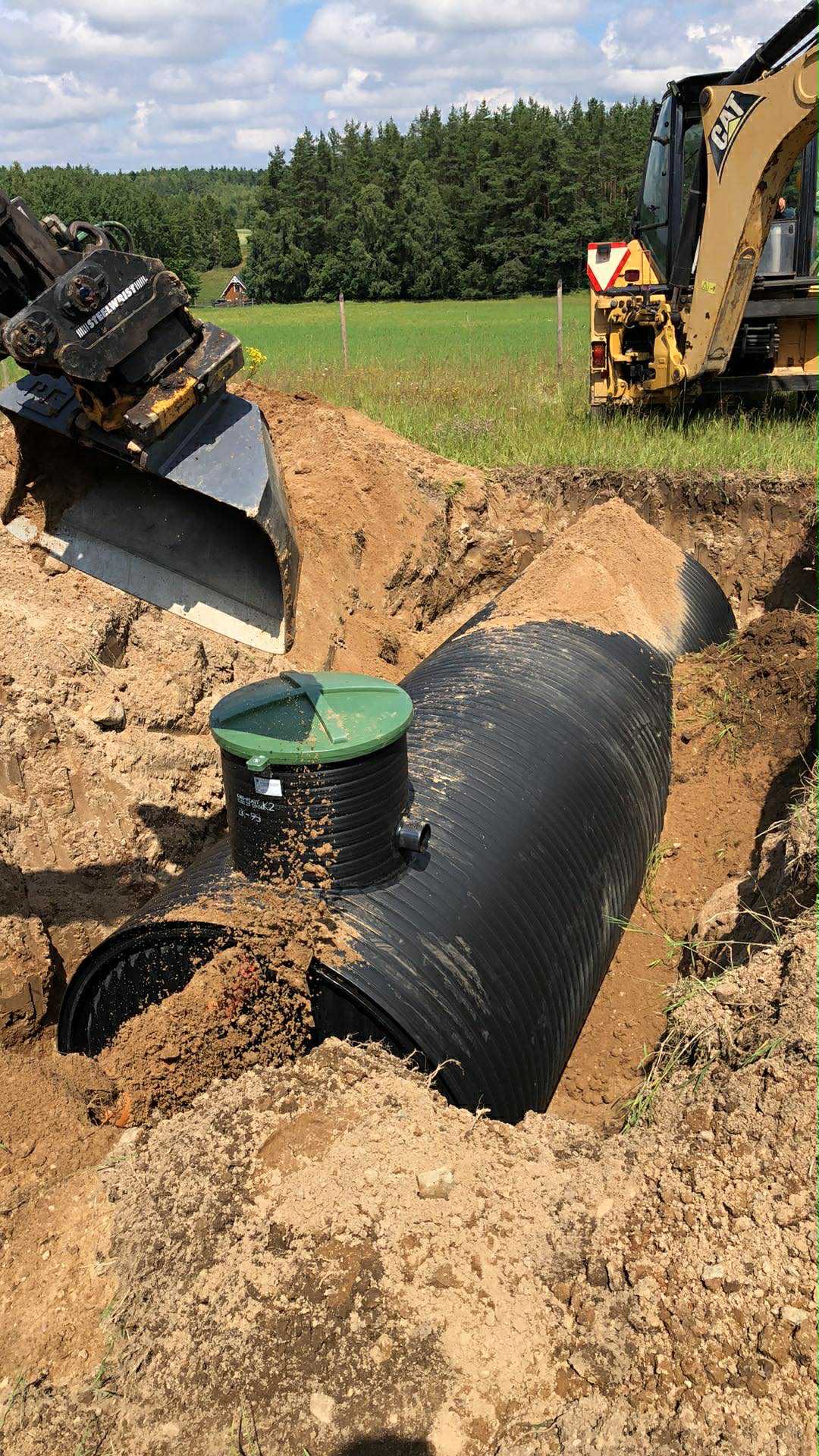 Montaż przydomowej oczyszczalni: czarna rura w wykopie, zielona pokrywa studzienki, koparka CAT w tle, słoneczny dzień.
