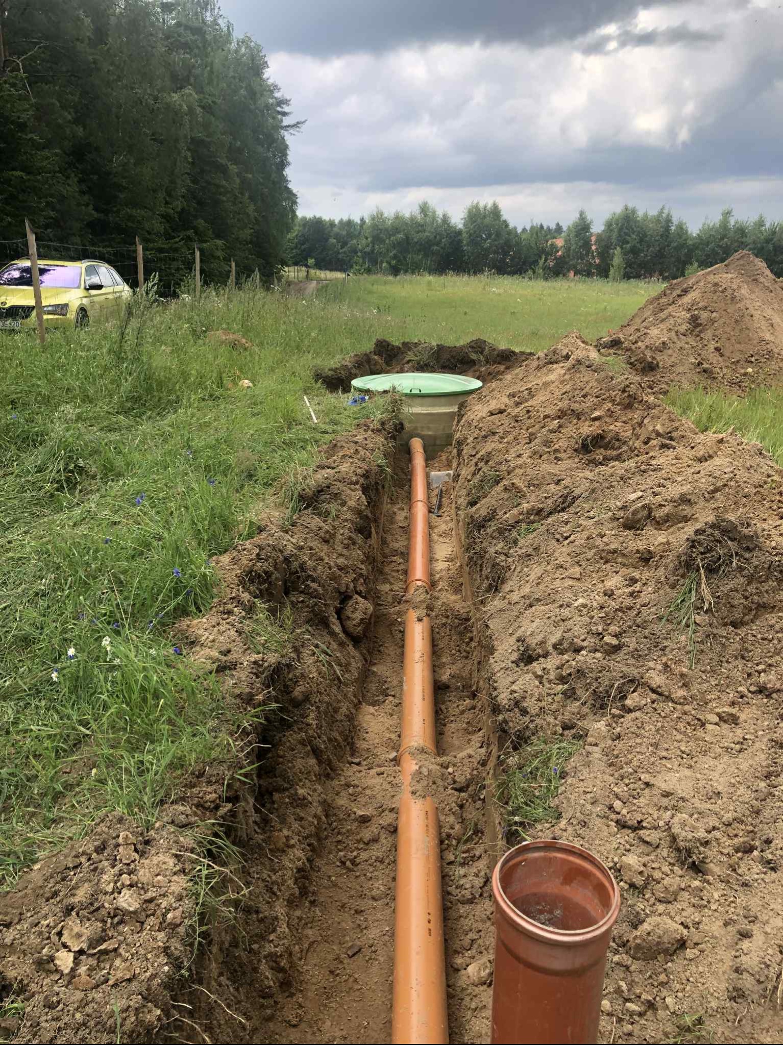 Montaż rur kanalizacyjnych w wykopie na tle zieleni i pochmurnego nieba. Widoczny fragment studzienki i żółty samochód w oddali.