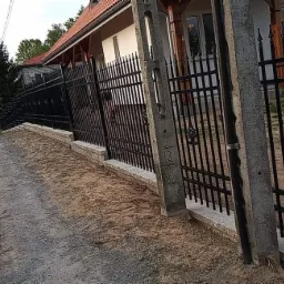 Czarne, metalowe ogrodzenie posesji z bramą i furtką, osadzone na niskim murku z jasnej kostki, widok z drogi gruntowej.