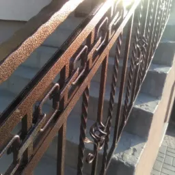 Metalowa balustrada schodowa z dekoracyjnymi elementami, pomalowana na brązowo, uchwycona pod kątem z mocnym światłem słonecznym.