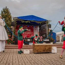 Uliczny występ z udziałem elfa żonglującego diabolo, elfa wskazującego palcem, elfa pochylonego nad skrzynią, kobiety na szczudłach w białym stroju i Świętego Mikołaja na linie, na tle sceny...