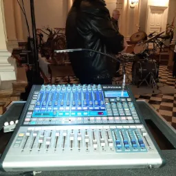 Profesjonalny cyfrowy mikser audio marki PreSonus z podświetlanymi suwakami i pokrętłami, ustawiony w kościele podczas przygotowań do wydarzenia, z widoczną perkusją w tle.