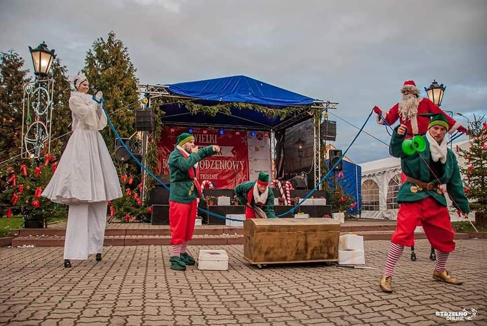 Uliczny występ z udziałem elfa żonglującego diabolo, elfa wskazującego palcem, elfa pochylonego nad skrzynią, kobiety na szczudłach w białym stroju i Świętego Mikołaja na linie, na tle sceny...
