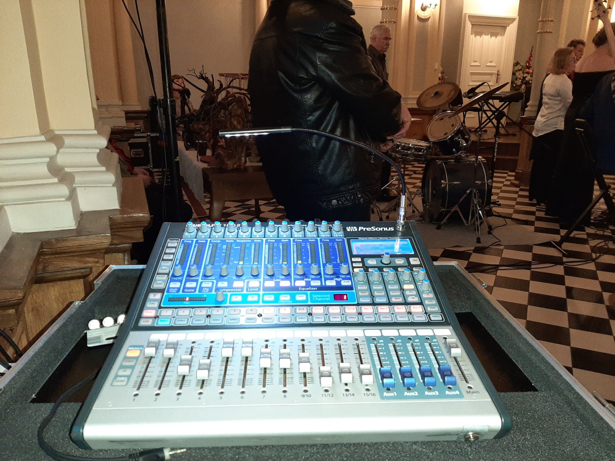 Profesjonalny cyfrowy mikser audio marki PreSonus z podświetlanymi suwakami i pokrętłami, ustawiony w kościele podczas przygotowań do wydarzenia, z widoczną perkusją w tle.