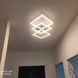 Piotr Kozak - Nowoczesna lampa sufitowa LED, składająca się z trzech kwadratowych ram, emituje ciepłe światło w nowym wnętrzu, gdzie widać przygotowane otwory na drzwi.