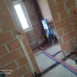 Surowe wnętrze w trakcie instalacji: widoczne przewody elektryczne w peszlach, rury CO i wodne, oraz stelaż podtynkowy WC Geberit przy oknie.