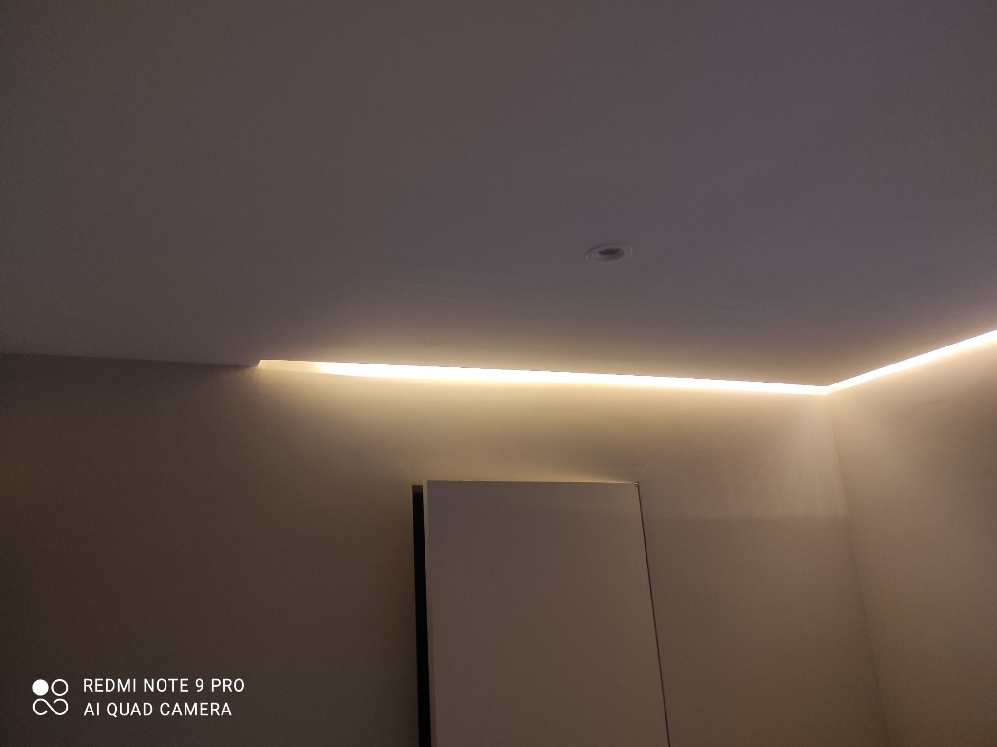 Nowoczesne oświetlenie LED w suficie podwieszanym, ciepłe światło, minimalistyczny design, widoczny fragment białej szafki, oświetlenie punktowe.