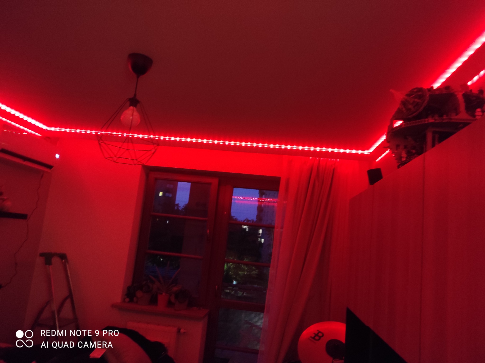 Pokój oświetlony intensywnym czerwonym światłem z taśmy LED zamontowanej pod sufitem, z widoczną lampą wiszącą i oknem z firanką w tle.