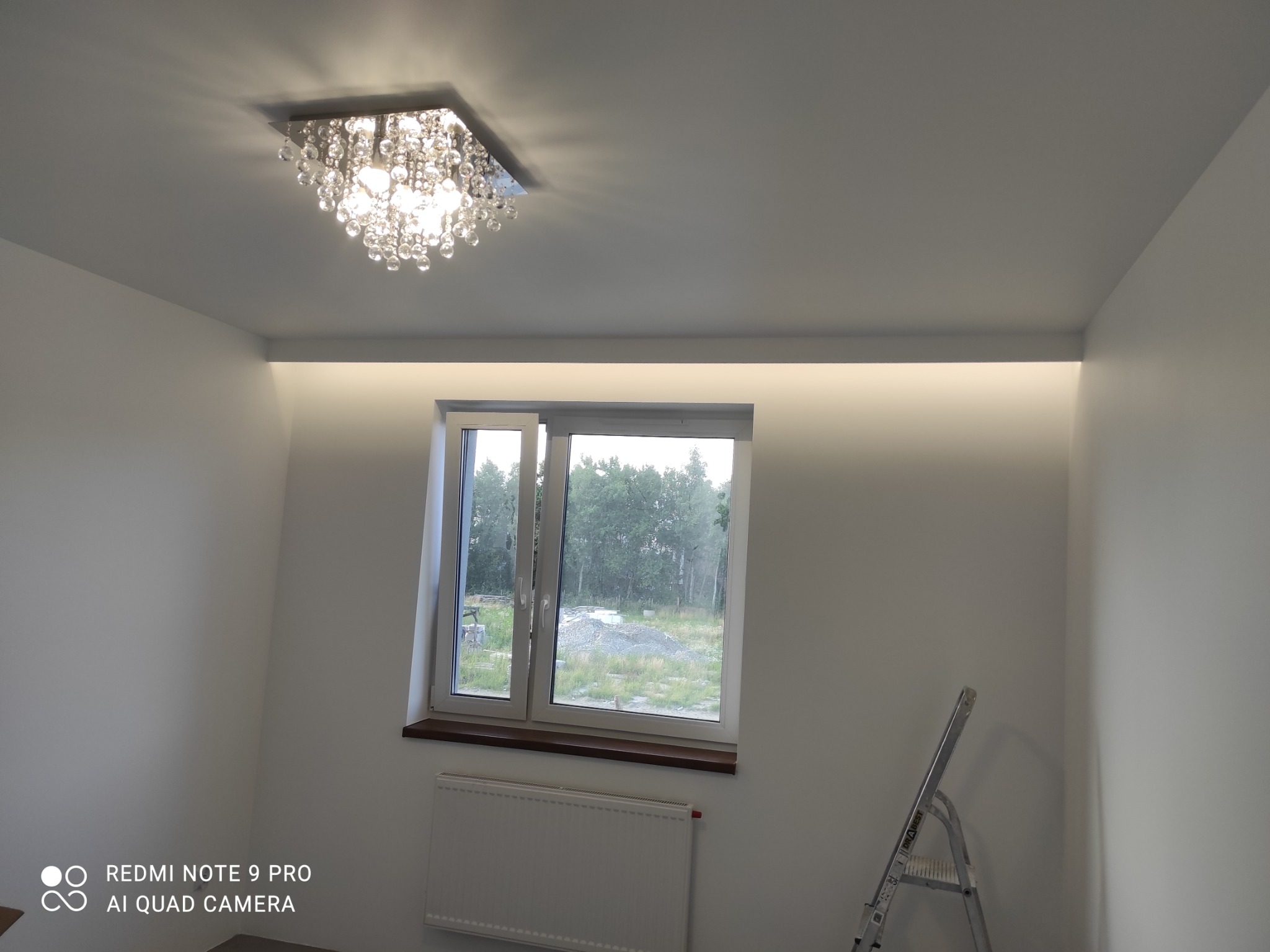 Kryształowy żyrandol sufitowy w jasnym pokoju z oknem i podświetleniem LED w suficie podwieszanym. Aluminiowa drabina oparta o ścianę.