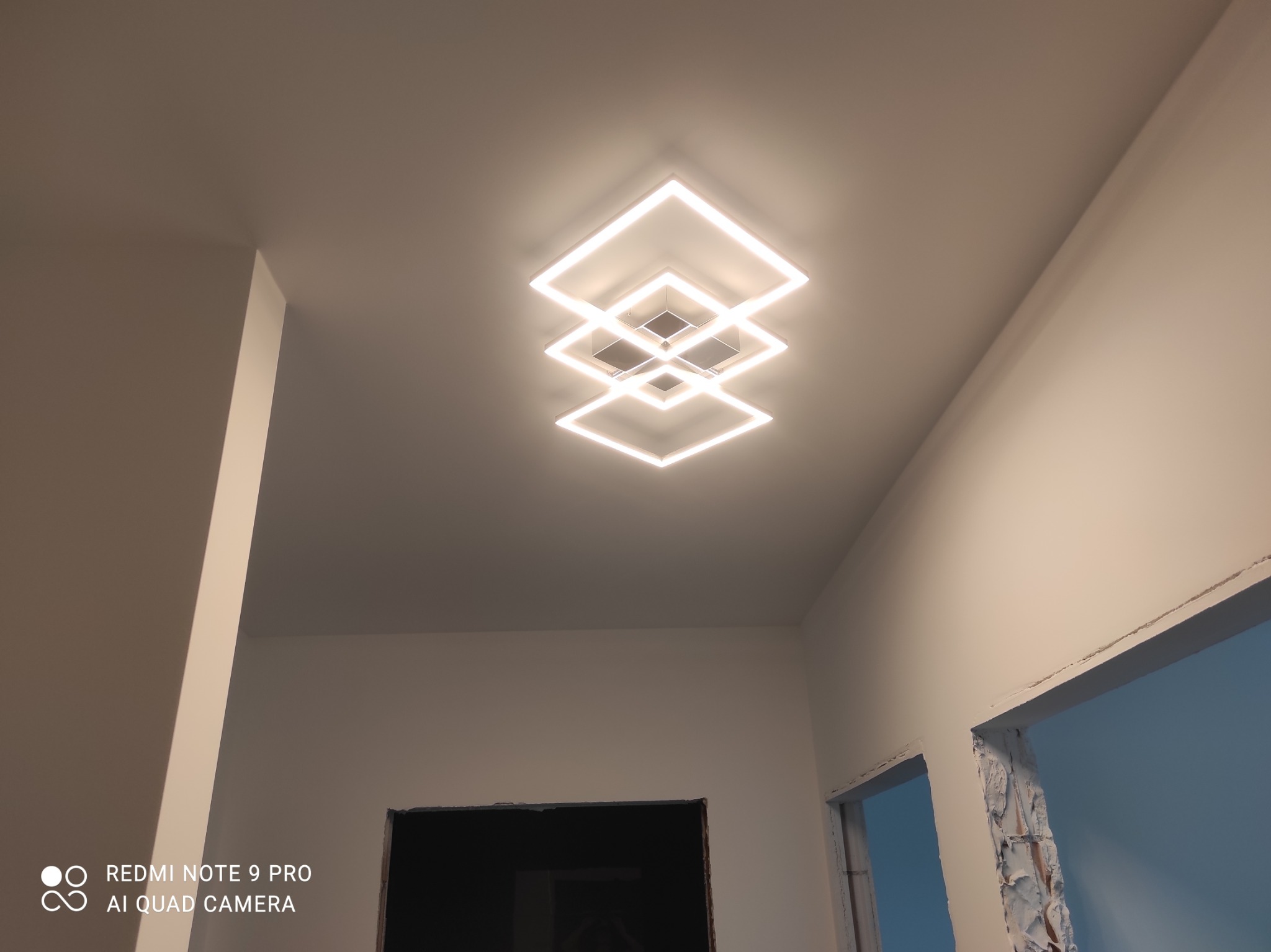 Nowoczesna lampa sufitowa LED, składająca się z trzech kwadratowych ram, emituje ciepłe światło w nowym wnętrzu, gdzie widać przygotowane otwory na drzwi.