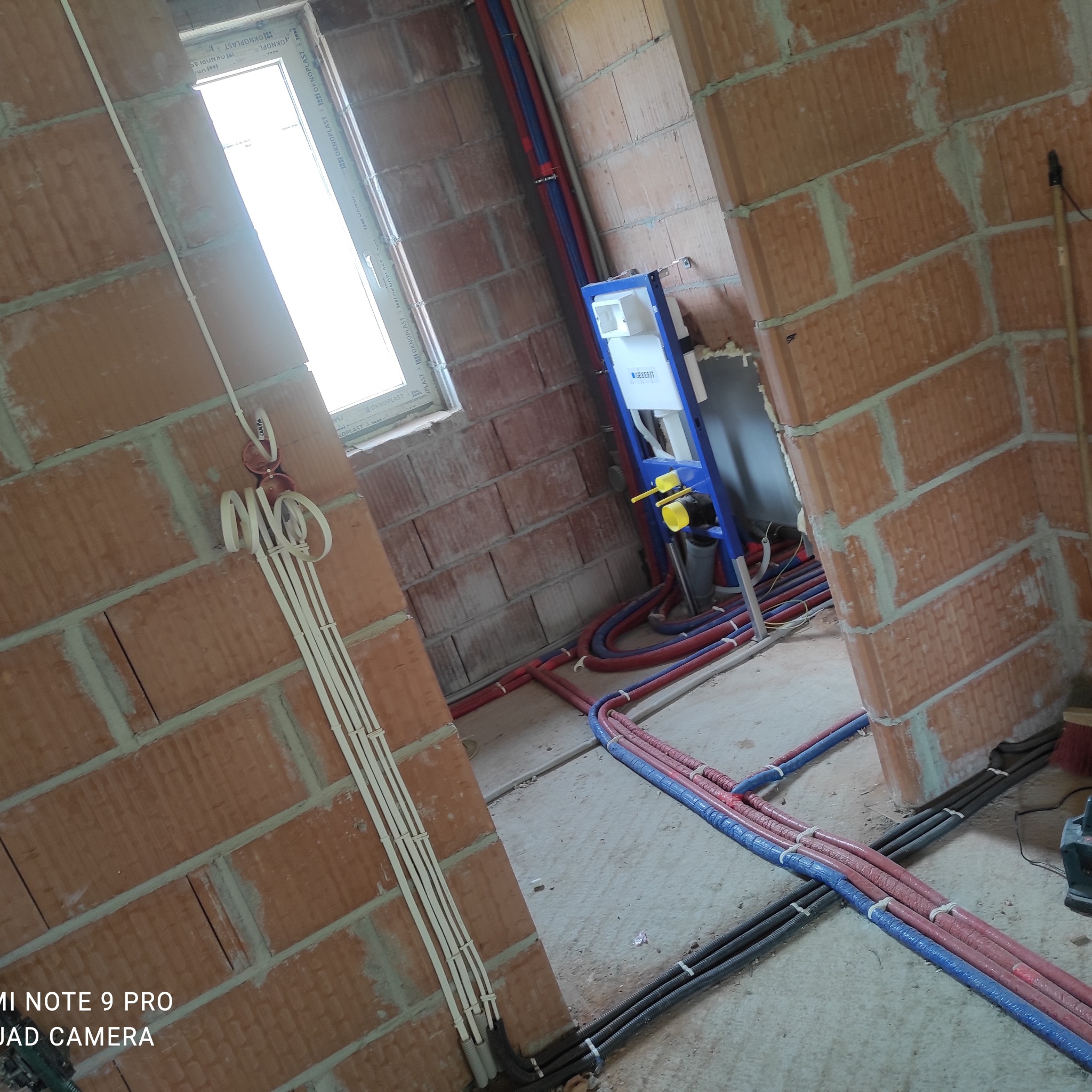 Surowe wnętrze w trakcie instalacji: widoczne przewody elektryczne w peszlach, rury CO i wodne, oraz stelaż podtynkowy WC Geberit przy oknie.