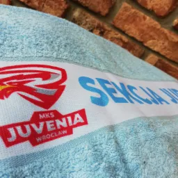 Jasnoniebieski ręcznik frotte z nadrukowanym logo klubu sportowego MKS Juvenia Wrocław i napisem 'Sekcja Judo', leżący na tle ściany z czerwonej cegły.