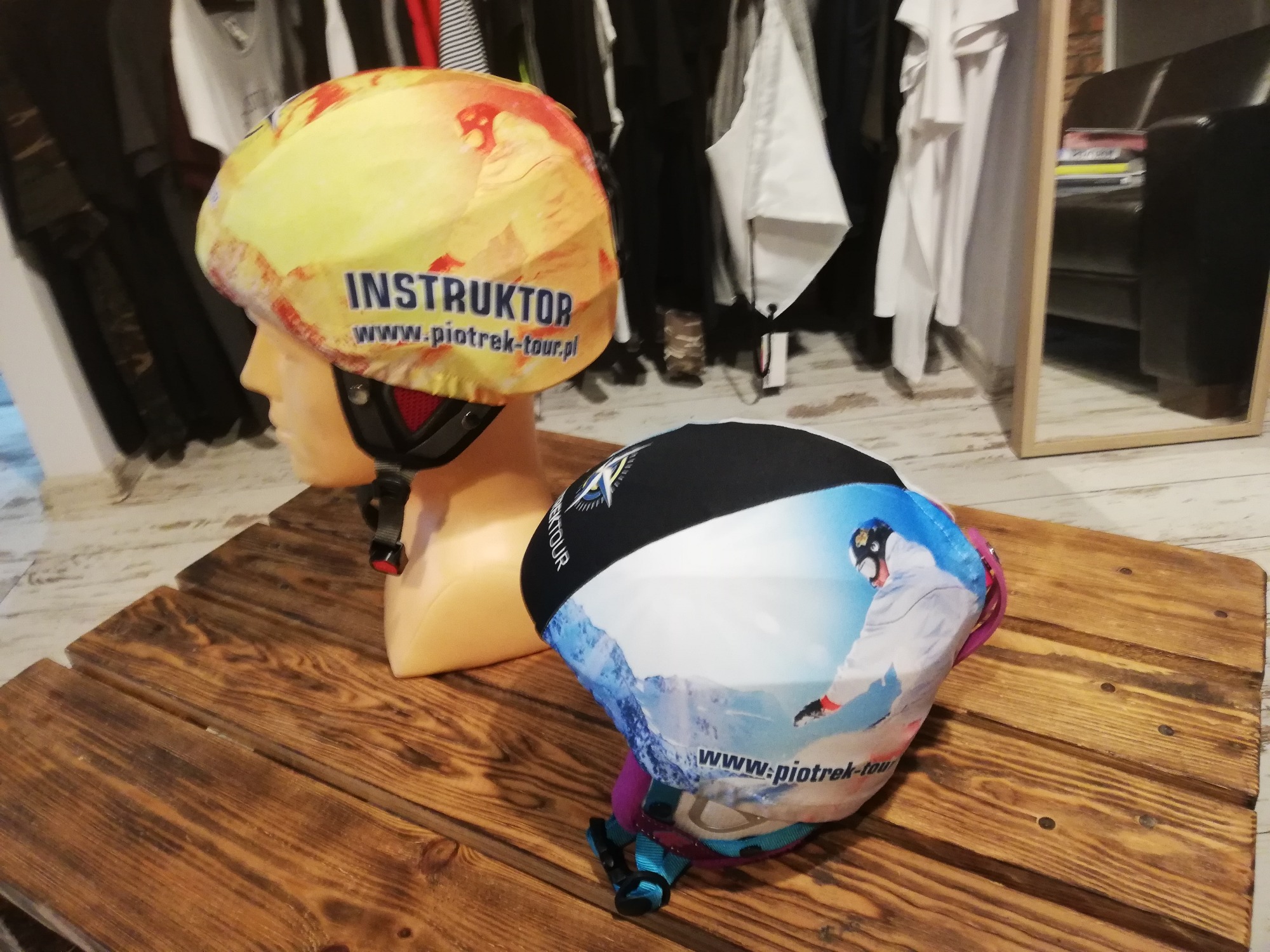Dwa kaski narciarskie z nadrukami leżące na drewnianym stole, jeden z napisem 'INSTRUKTOR' i adresem strony, drugi z grafiką przedstawiającą narciarza.