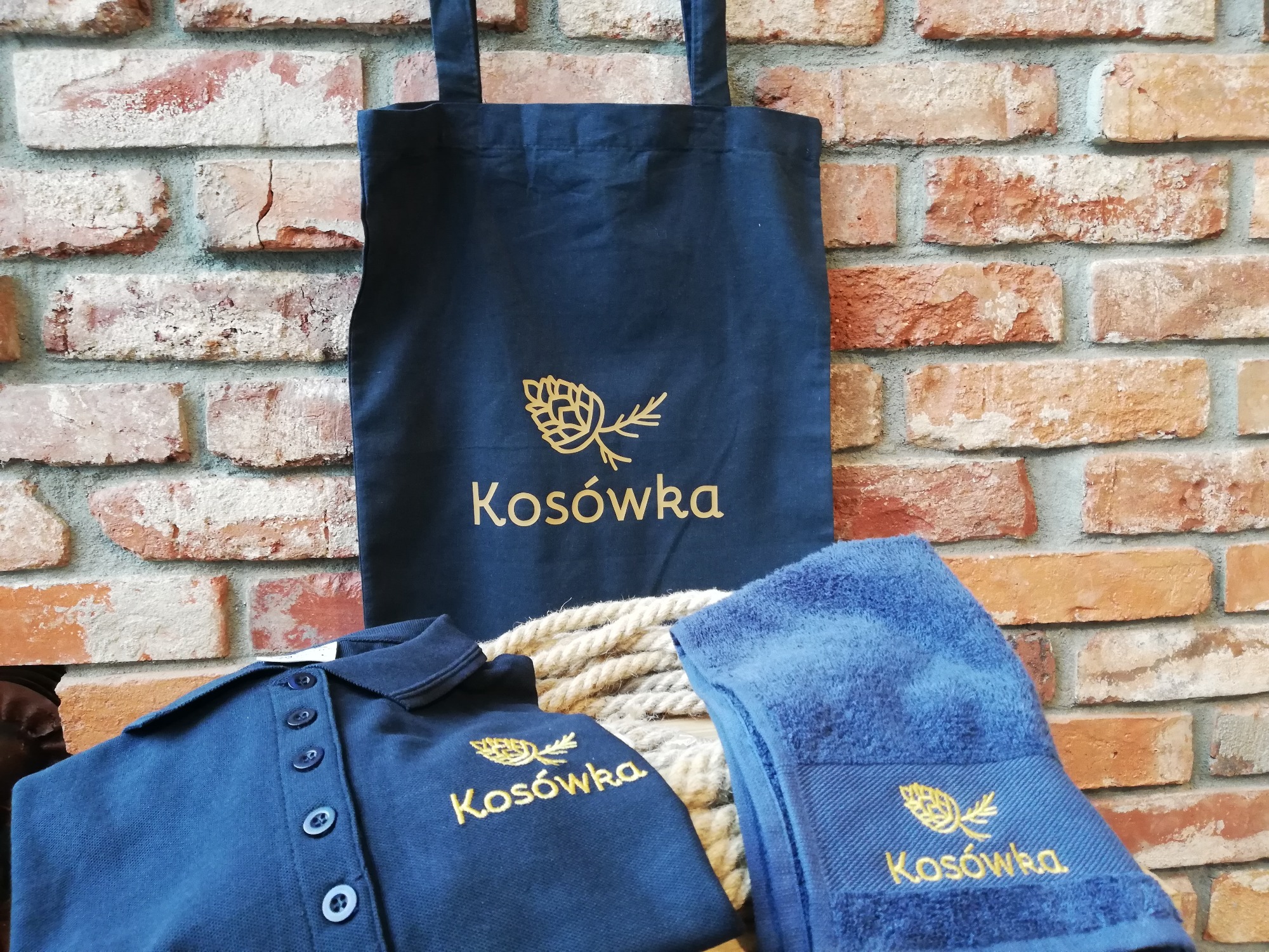 Granatowa torba bawełniana, koszulka polo i ręcznik z haftowanym złotym logo i nazwą 'Kosówka' na tle ceglanej ściany i sznura.