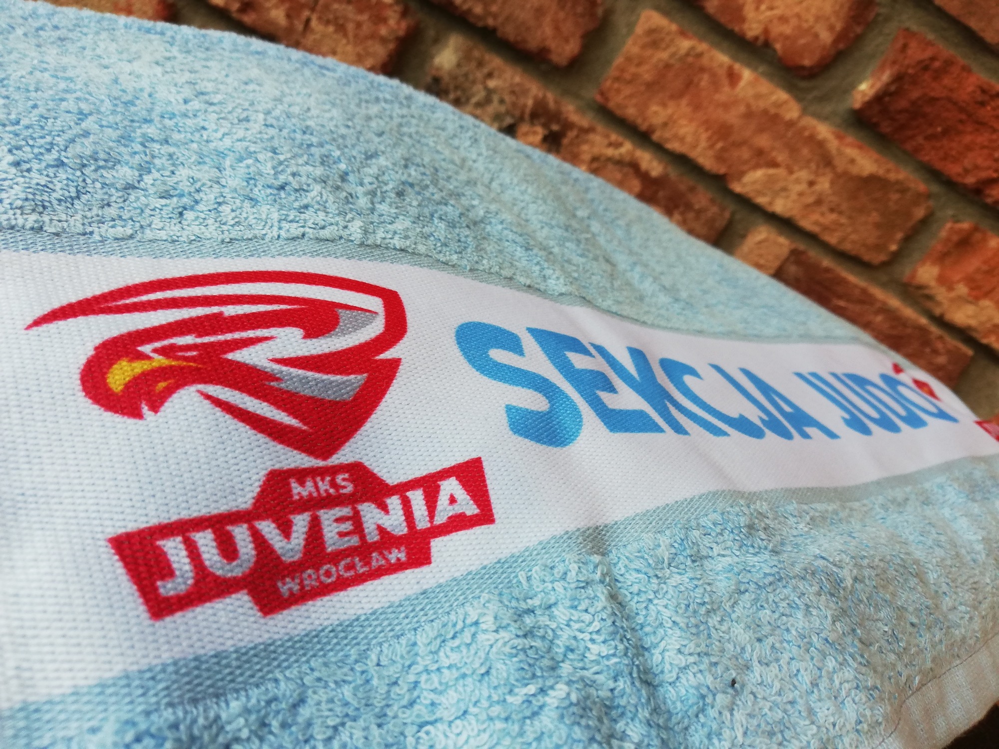 Jasnoniebieski ręcznik frotte z nadrukowanym logo klubu sportowego MKS Juvenia Wrocław i napisem 'Sekcja Judo', leżący na tle ściany z czerwonej cegły.