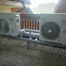 Dwie jednostki zewnętrzne pompy ciepła Unical HP.OWER full inverter, zamontowane na metalowej konstrukcji na żwirowym podłożu, widok z boku z drewnianą balustradą i dachem w tle.