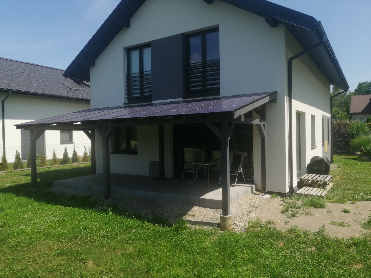 Zadaszenie tarasu z paneli fotowoltaicznych bifacjalnych, widok na nowoczesny dom z szarymi oknami i zielony trawnik.