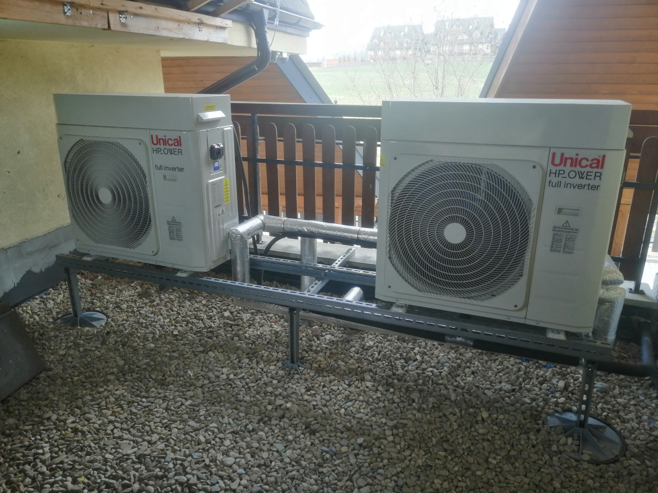 Dwie jednostki zewnętrzne pompy ciepła Unical HP.OWER full inverter, zamontowane na metalowej konstrukcji na żwirowym podłożu, widok z boku z drewnianą balustradą i dachem w tle.