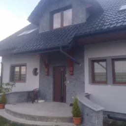 Dwukondygnacyjny dom jednorodzinny z szarą elewacją imitującą kamień, brązowymi ramami okiennymi i ciemną dachówką, wejście z półokrągłym tarasem i małą architekturą ogrodową.