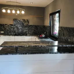 Kuchnia z blatem z granitu Juparana, widoczne białe szafki, płyta grzewcza, okno z kwiatami i nowoczesne oświetlenie sufitowe.