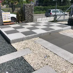 Wystawa zewnętrzna z szachownicą z jasnoszarych i ciemnoszarych płyt granitowych, otoczona żwirem i kamiennymi gabionami w tle.