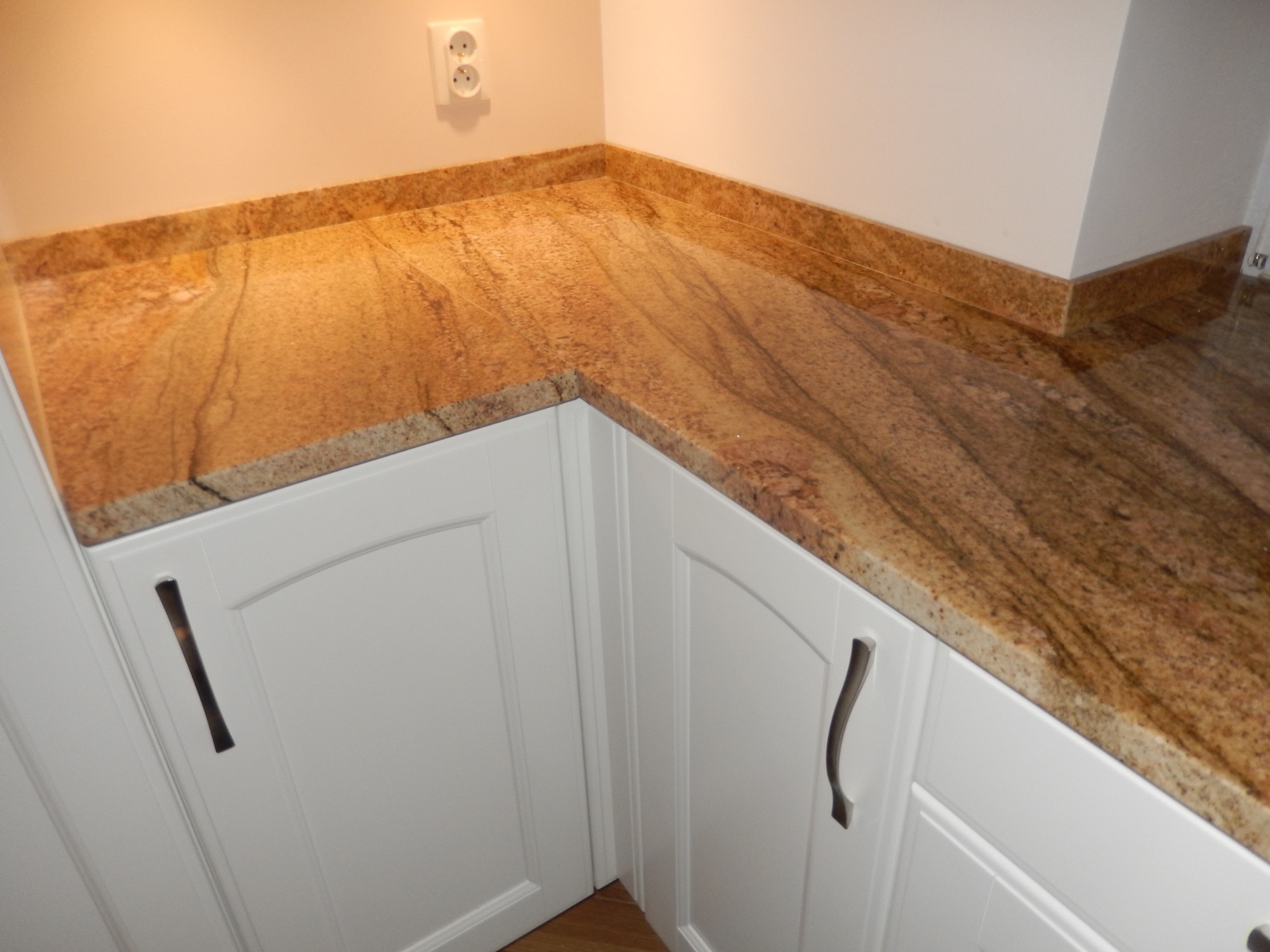 Kuchenny blat z granitu Imperial Gold, zamontowany na białych szafkach z frezowanymi frontami i metalowymi uchwytami, widoczny fragment ściany z gniazdkiem elektrycznym.