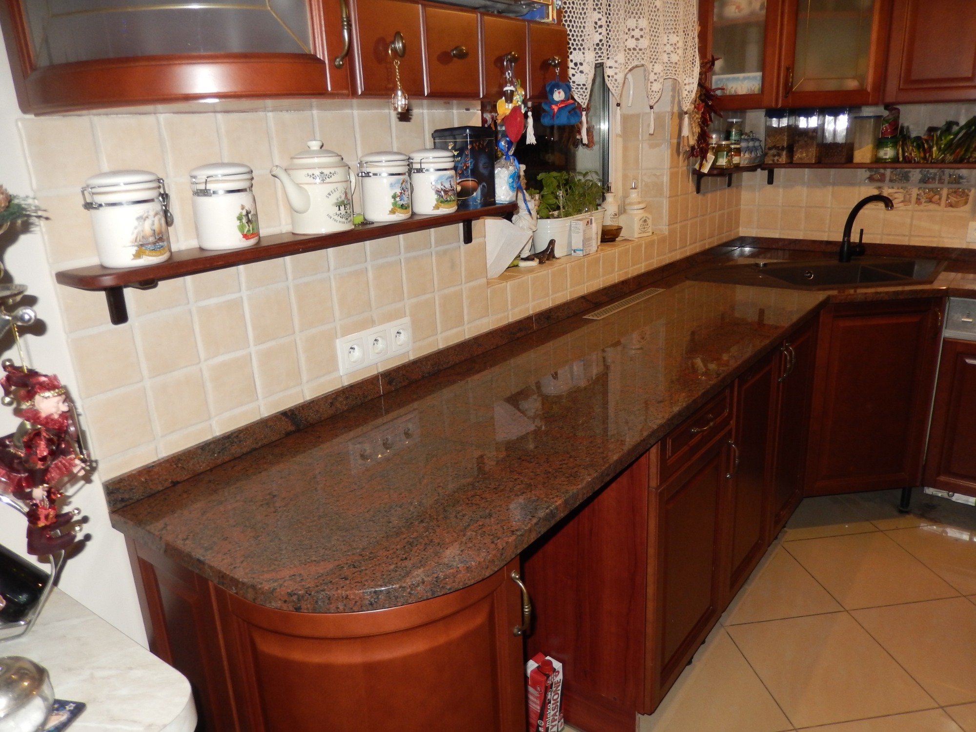 Kuchenny blat z granitu Multicolor Red, z zaokrąglonym narożnikiem, zamontowany na ciemnobrązowych szafkach, widoczne gniazdka elektryczne w ścianie nad blatem.