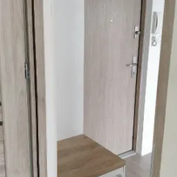 Wnęka w przedpokoju z siedziskiem i szafką na buty, obok drzwi wejściowe z wizjerem i domofonem.