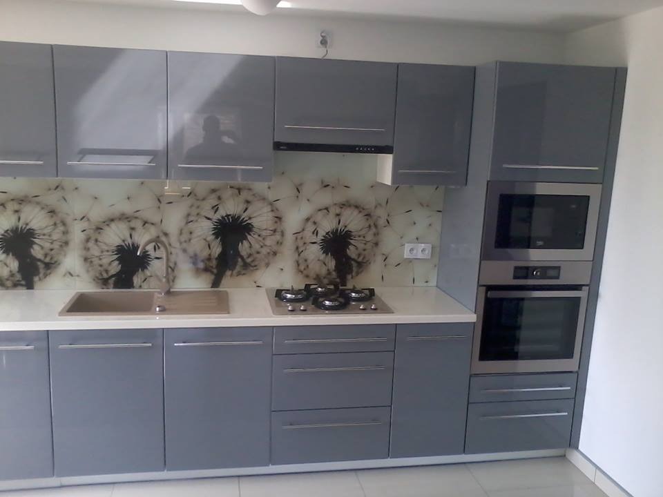 Szara zabudowa kuchenna z frontami na wysoki połysk, blatem w kolorze złamanej bieli i panelem szklanym z motywem dmuchawców. Widoczna płyta gazowa i piekarnik w zabudowie.