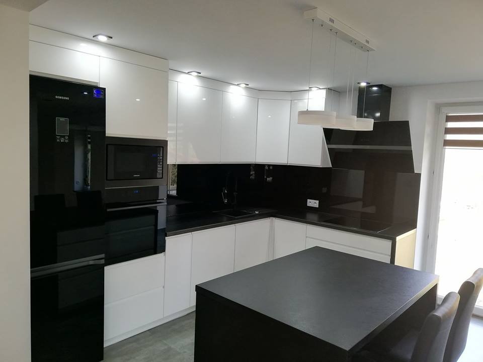 Nowoczesna kuchnia: białe szafki z połyskiem, czarny blat i sprzęt AGD, wyspa kuchenna z krzesłami, oświetlenie punktowe i wiszące lampy.