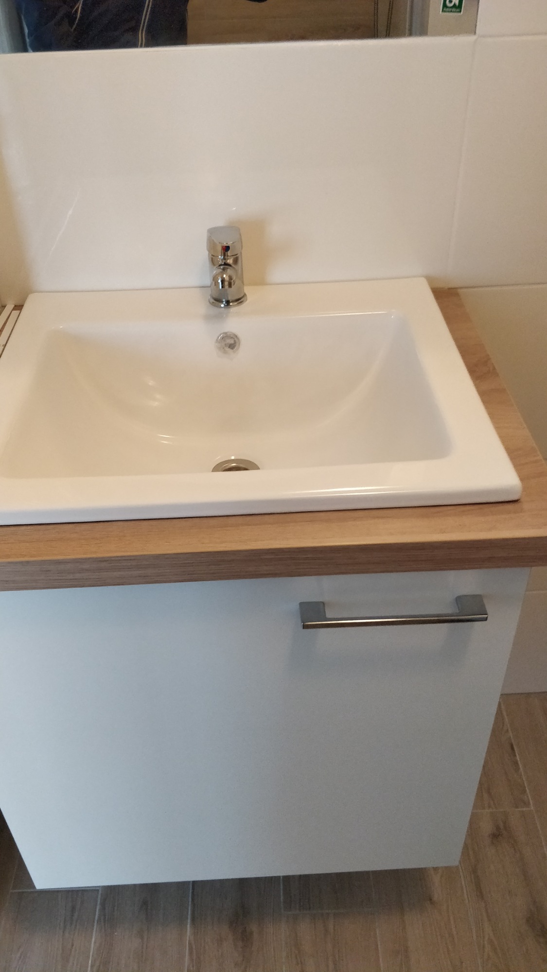 Biała umywalka nablatowa z chromowaną baterią, osadzona na szafce z drewnopodobnym blatem, widoczna część lustra w tle.