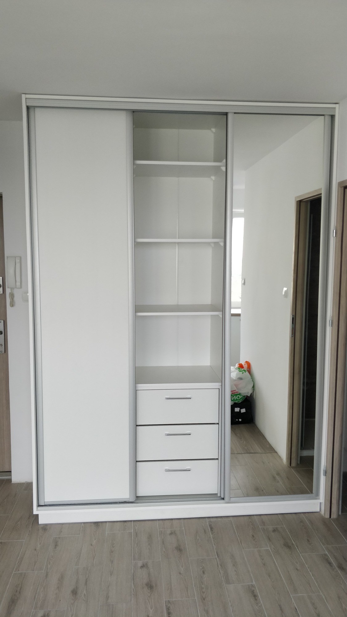 Biała szafa przesuwna z lustrem w przedpokoju, widoczne półki i szuflady, na podłodze panele imitujące drewno.