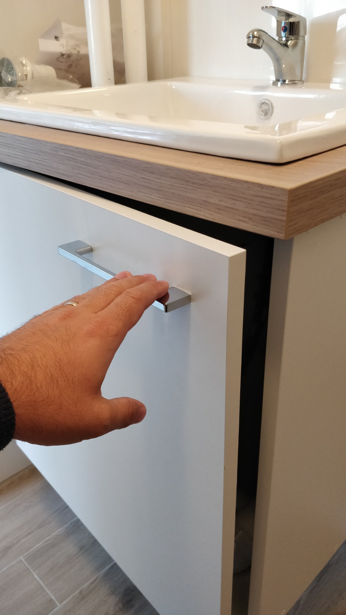 Otwarta biała szafka pod umywalką z drewnianym blatem, widoczna dłoń z obrączką na uchwycie.