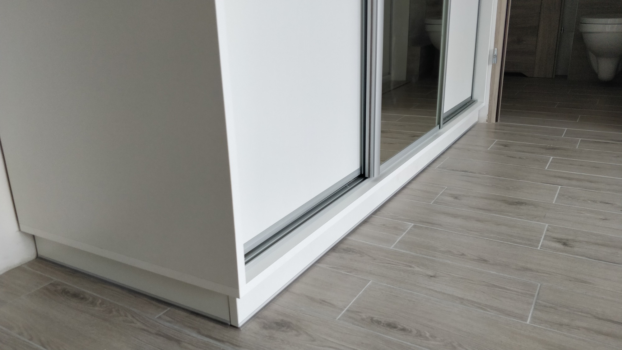 Biała szafa przesuwna z lustrzanymi drzwiami, widok z dołu, na podłodze z płytek imitujących drewno, z widoczną toaletą w odbiciu.