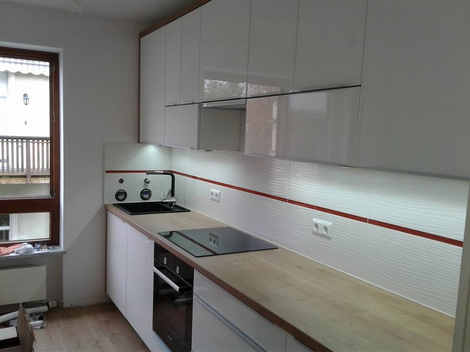 Nowoczesna kuchnia z białymi, błyszczącymi szafkami, drewnianym blatem i falistymi płytkami z czerwonym akcentem, widok na okno z balkonem.