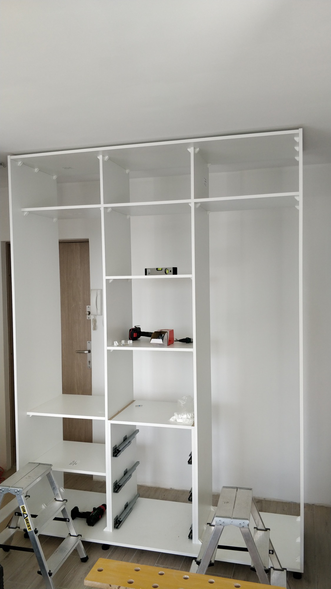 Montaż białej szafy wnękowej z widocznymi prowadnicami szuflad, poziomica i wkrętarka na półce, drabiny monterskie po bokach.