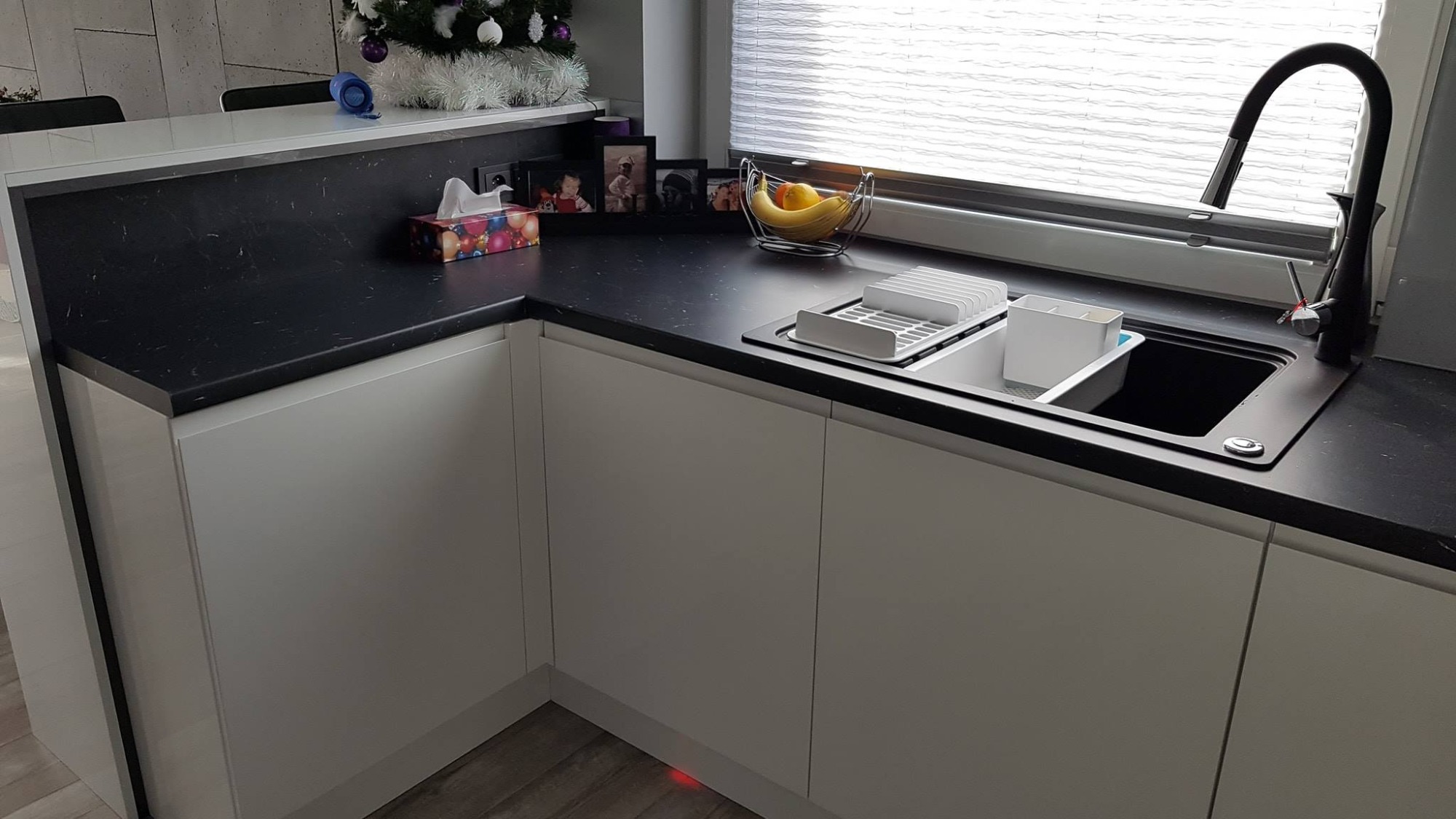 Nowoczesna kuchnia z czarnym blatem i białymi szafkami, widoczny zlew, bateria i ociekacz, w tle okno z żaluzjami i dekoracja świąteczna.