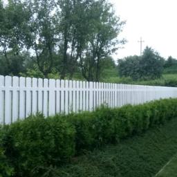 Trawka - Długie, białe, drewniane ogrodzenie panelowe z zaokrąglonymi szczytami, oddzielające ogród od zielonego wzgórza, z niskimi krzewami posadzonymi wzdłuż jego podstawy.