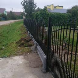 Trawka - Nowoczesne, czarne ogrodzenie metalowe z betonową podmurówką, widok z boku, na tle zieleni i domów jednorodzinnych w pochmurny dzień.