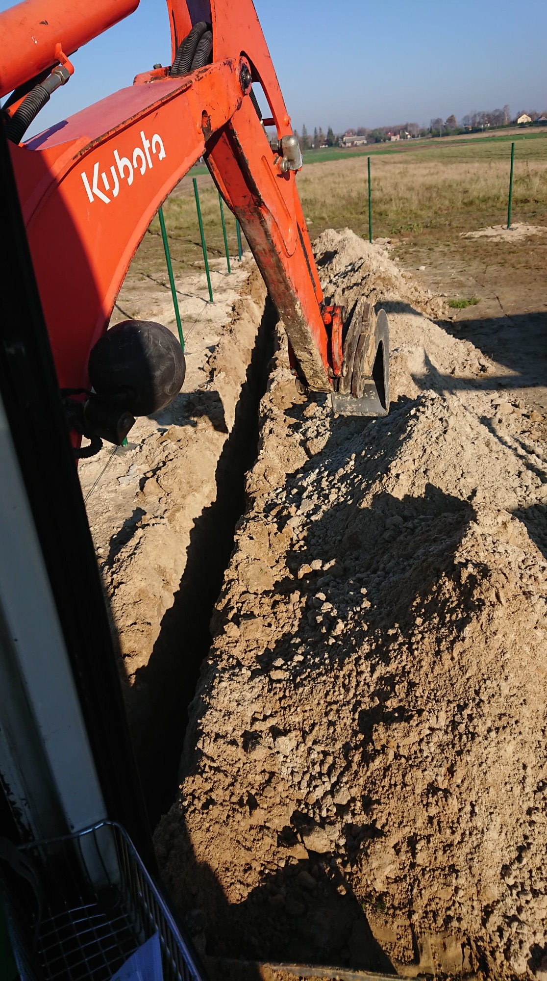 Pomarańczowa koparka Kubota w trakcie wykonywania wąskiego wykopu w piaszczystej glebie, widoczne zielone słupki ogrodzeniowe w tle.