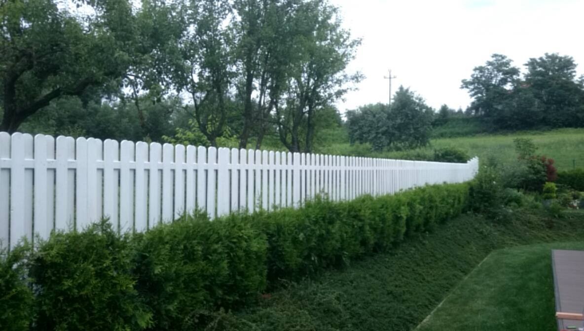 Długie, białe, drewniane ogrodzenie panelowe z zaokrąglonymi szczytami, oddzielające ogród od zielonego wzgórza, z niskimi krzewami posadzonymi wzdłuż jego podstawy.