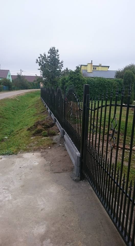 Nowoczesne, czarne ogrodzenie metalowe z betonową podmurówką, widok z boku, na tle zieleni i domów jednorodzinnych w pochmurny dzień.