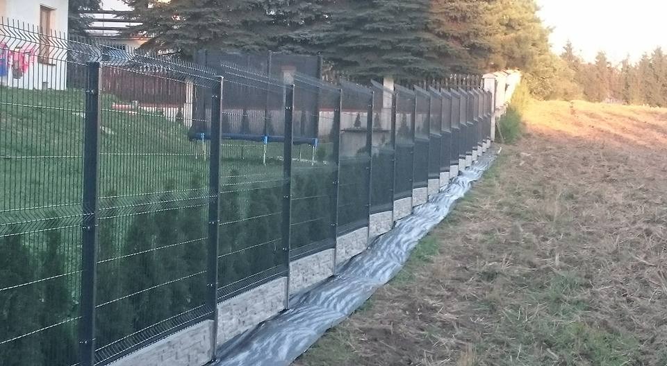 Długi fragment panelowego ogrodzenia z podmurówką betonową, zabezpieczony folią, oddzielający posesję od pola z uschniętą trawą.