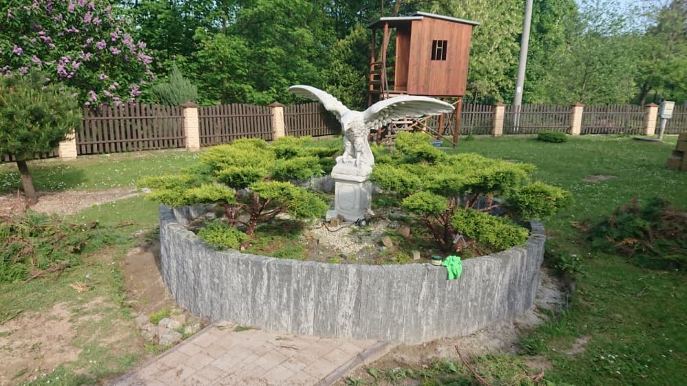 Ogród z okrągłą rabatą otoczoną betonowym murkiem, w centrum rzeźba orła na postumencie, otoczona karłowatymi drzewkami iglastymi, w tle drewniany domek na palach i drewniane ogrodzenie.